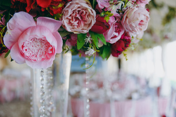 Beautiful decoration of the wedding banquet under the awning in pink, burgundy and white tones. High vase with a large flower arrangement, decorated with beads on the dining table in the banquet hall