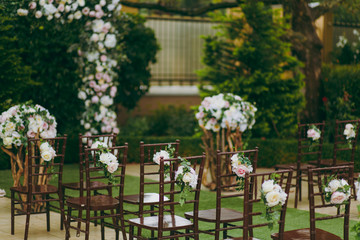 Fototapeta na wymiar Beautiful decoration of a wedding ceremony in a green autumn garden. Brown wooden chairs for guests on either side of the walkway, which leads to a festive arch decorated with flowers