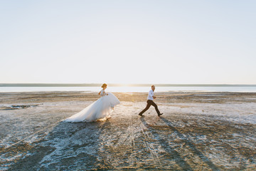 Beautiful wedding photosession. Handsome unshaved groom in a black trousers and young cute bride in white lace pattern dress with exquisite hairstyle on walk along the coastline near the sea sunset
