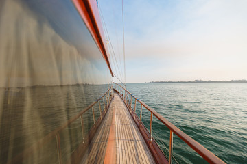 Obraz premium The curtained window on the wooden deck of brown yacht with wooden rails and golden details in azure wavy sea near the coastline against the backdrop of clear blue sky, which are reflected in window