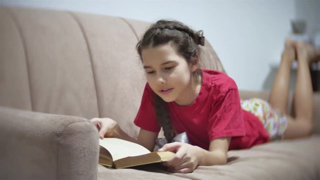 girl teen reading a book lying on sofa indoors. girl kid reading a book lying on a indoors sofa