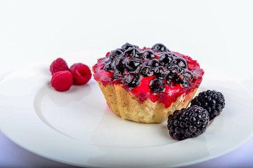 cake in the form of a basket of shortbread cookies, filled with black currant, filled with a shiny red jelly, lies on a white saucer and is decorated with berries of raspberry and blackberries