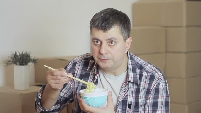 A middle-aged man sitting on the floor in a room with cardboard boxes and eating noodles with Chinese sticks looking at the TV