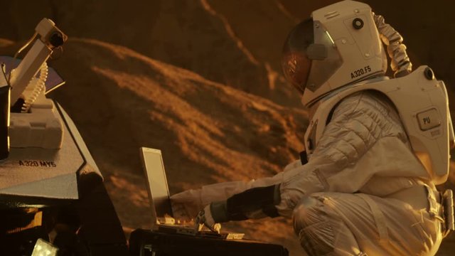 Astronaut in the Space Suit Works on Laptop, Adjusting Rover on a New Alien Red Planet, Presumably Mars. Day Light High-Tech Space Exploration. Shot on RED EPIC-W 8K Helium Cinema Camera.