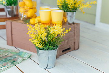 A bouquet of yellow flowers in a vase in the form of a bucket on the background of a wooden box with yellow glasses and lemons.