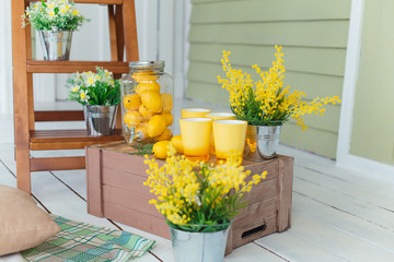 Lemons in a glass jar and next to yellow glasses and bouquets of yellow flowers in vases in the form of a bucket on a wooden brown box. Spring decoration. - Powered by Adobe