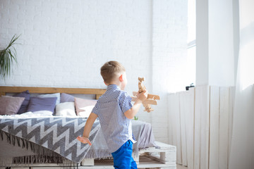 A little boy runing around the room and playing with a wooden toy plane.