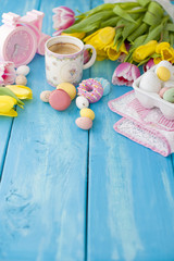Easter eggs of different colors. A pink alarm clock and a cup of coffee, coffees and a macaroni for breakfast. Blue wooden background and spring tulips. A pink napkin. Free space for text