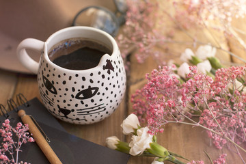 Aramatic coffee in a mug. Notepad is black. Decor of pink and white flowers on the table. Free space for text. Card. A view from above.