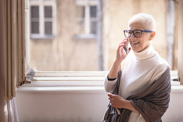 Beautiful senior lady talking on her cell phone, standing next to a big home window, enjoying her...