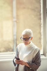 Beautiful senior lady typing a message on her cell phone, standing next to a big home window, enjoying her chat