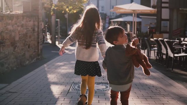 Little girl and boy walk together holding hands. Slow motion. Back view. Two kids wander around old town on a sunny day.