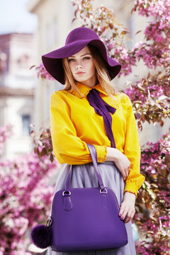 Outdoor portrait of young beautiful girl posing in street with blooming trees, wearing stylish hat, yellow shirt, tulle skirt, holding violet handbag. City lifestyle. Female spring fashion concept

