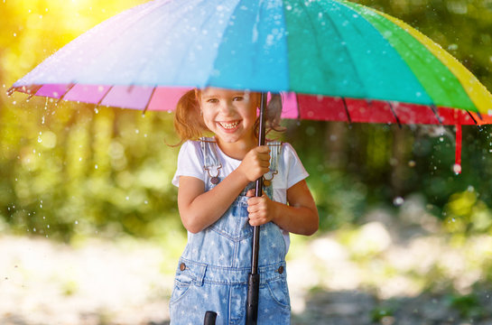Happy child girl laughs and plays under summer rain with an umbrella.