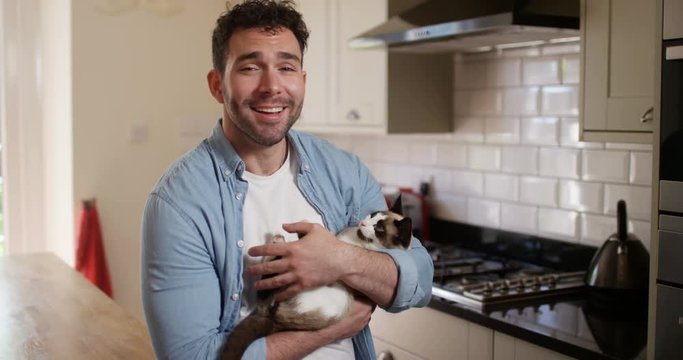 4K Cheerful man holding annoyed pet cat making a video call in the kitchen at home