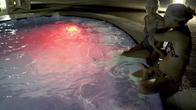 Backlit Shot At Night Of Two Women Sitting On The Edge And Dangling Their Legs In A Large Bubbling Hot Tub.