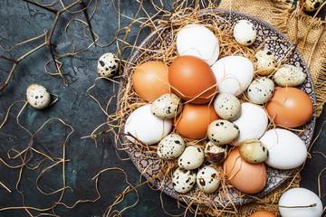 Multicolored chicken and quail eggs with straw and branches, spring Easter composition, black background, top view