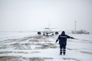 Member of ground crew park passenger airliner on airport apron in blizzard. passenger airliner taxi at airport in snow. Modern twin-engine passenger airplane taxiing at airport during snow blizzard