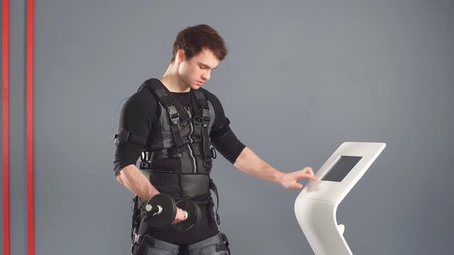 Man in Electrical Muscular Stimulation suit standing with dumbbells near ems tablet and push on screen.