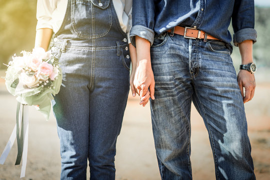 Young loving couple holding hands each other with wedding bouquet flowers , Bride and groom in vintage jeans standing together in soft light of sunset