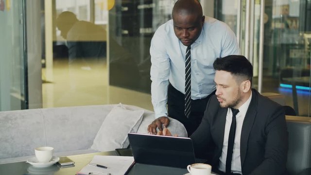 Two business partners discussing actively their startup project sitting in stylish cafe. Bearded businessman sitting ang talking his colleague while looking at laptop computer