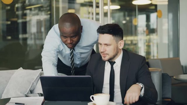 Two business partners discussing actively their startup project sitting in stylish cafe. Bearded businessman sitting ang talking his colleague while looking at laptop computer
