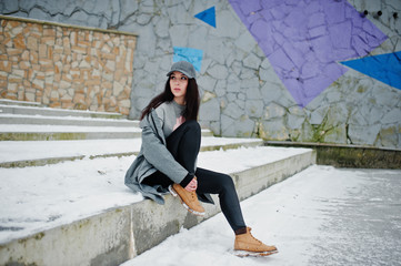 Stylish brunette girl in gray cap, casual street style on winter day against colored wall.