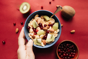 female hand holding bowl with delicious crispy cornflakes with kiwi pieces and cranberries,  healthy breakfast on red background