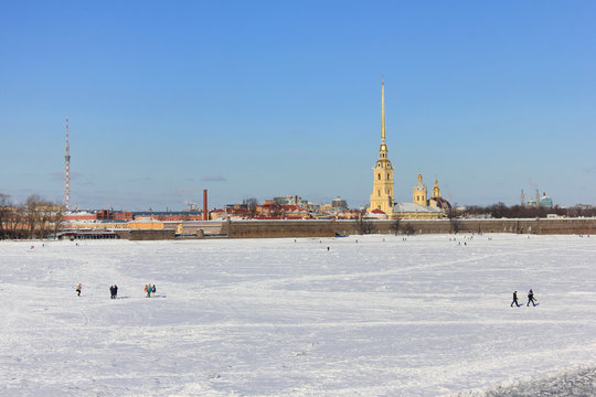 People Crossing Frozen River in Downtown Saint-Petersburg, Russia. Winter Scene Cityscape View of Peter and Paul Fortress and Neva River Covered in Ice with People Walking Across on Cold Winter Day.