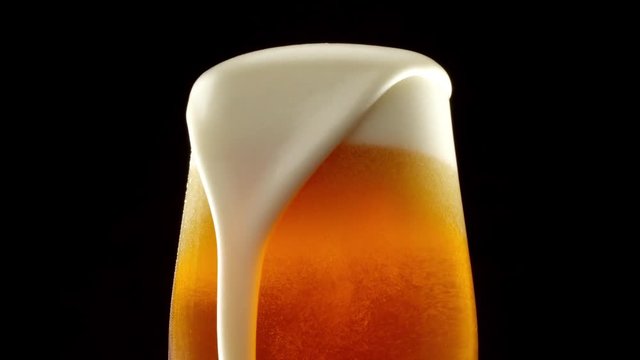 Stream of beer pours into a rotating beer glass. Close up 4K video. Black background.