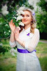 Obraz premium Beautiful blonde young girl in white dress with purple artificial butterflies in hands against blooming white and purple lilac