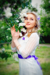 Obraz premium Beautiful blonde young girl in white dress with purple artificial butterflies in hands against blooming white and purple lilac