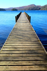 long wooden symetrical beautiful wooden jetty, jutting out from the centre of the image into a calm blue lake with hills of forest and meadows in background