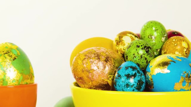 Close-up of Easter beautiful colorful eggs. Eggs lie in a yellow bowl, stand in stands, white background behind. A beautiful Easter composition made of eggs. Move the camera from left to right.