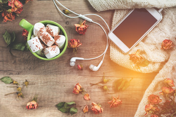 Cup of hot chocolate with marshmallow, white knitting wool, dried roses flowers, mobile phone and...