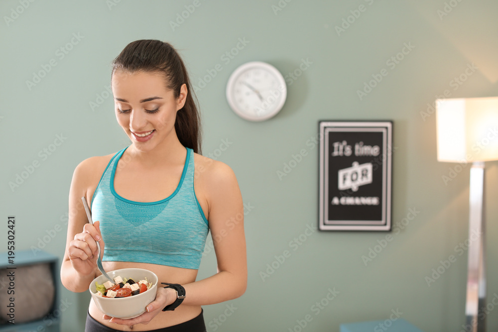 Poster Sporty young woman eating salad after fitness training at home