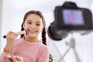 Cheerful mood. Pleasant upbeat girl smiling happily and applying powder to her cheek while filming...