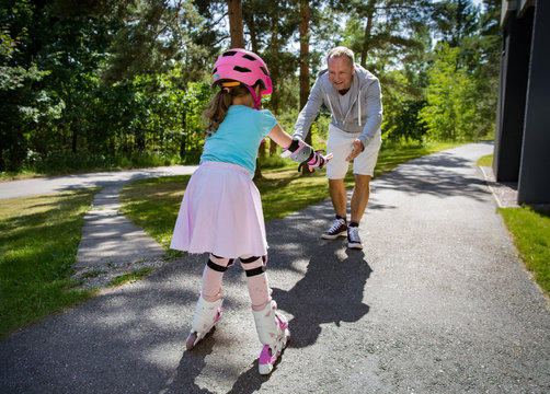 Father teaching his daughter to skate on roller skates. Happy kid in helmet learning skating. Family spending time together. Sunny summer day on suburb street.