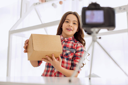 Popular video genre. Sweet pre-teen girl in a checked shirt holding a box in her hands and smiling cheerfully while recording an unboxing video on the camera
