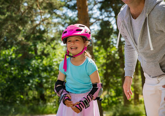 Father teaching his daughter to skate on roller skates. Happy kid in helmet learning skating. Family spending time together. Sunny summer day on suburb street.