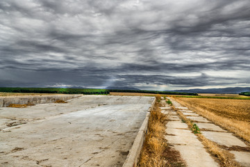 Fototapeta premium Summer landscape with storm clouds and wheat field after harvest. Sun rays over the field and forest. Storage place on field with dark sky.