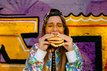 Cute fashionable young woman, enjoying eating a burger, closed her eyes. Dressed in stylish jacket and cap, in eyeglasses. Standing against the wall with graffiti.