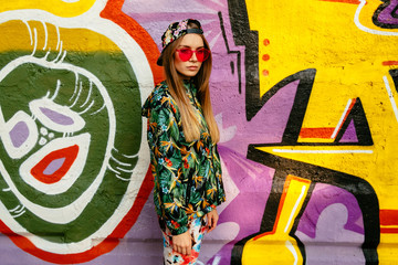 Trendy charming girl in colorful clothes, wearing stylish cap and red eyeglasses, standing with pensive face, posing near the wall with graffiti.