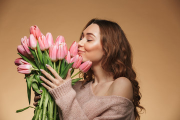 Image of lovely woman with long brown hair holding and smelling bouquet of pink flowers, isolated over beige background