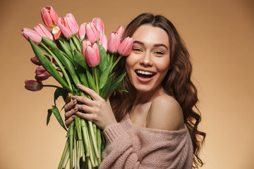 Cheerful young lady with long brown hair being excited to get bouquet of pink tulips on women's day, isolated over beige background