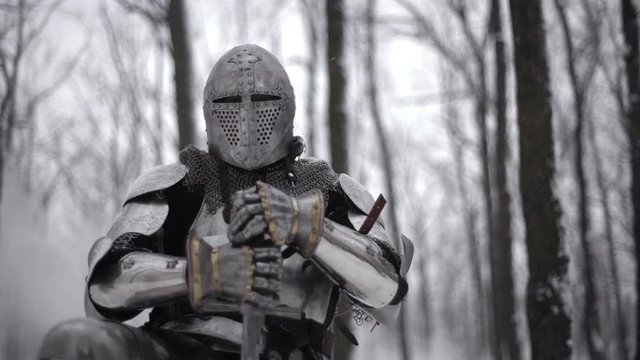 Portrait of medieval warrior putting on steel helmet while kneeling, and holding traditional sword in winter forest slow motion. Fantasy and reenactment
