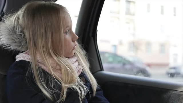 Sad child looking to the car window. Little blonde girl on a back seats in car.