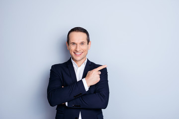 Attractive, corporate, positive, modern, fashionable man in formal wear with crossed hands pointing to copy space with forefinger, looking at camera, standing over gray background
