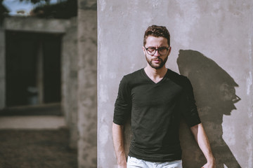 Obraz premium Angry handsome young bearded hipster guy in casual outfit and eyeglasses is leaning against the wall on a sunny day, with copy space zone on the left for your advertising, logo or text message