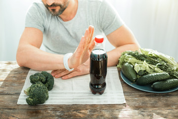 Bad idea. Unshaken concentrated decisive man refusing to drink cola gesticulating and sitting by the table.